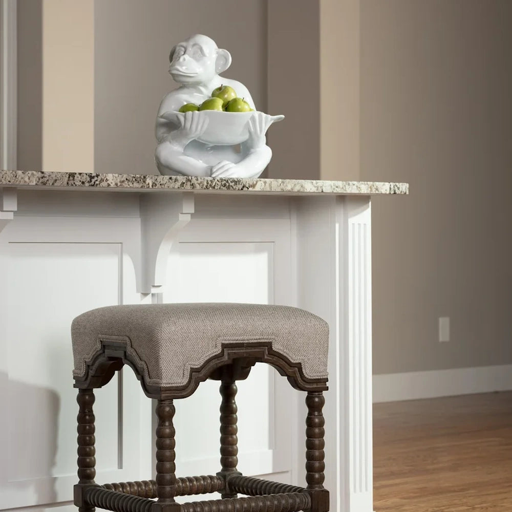 White ceramic monkey statue holding green apples on a granite kitchen island counter above a gray upholstered bar stool.