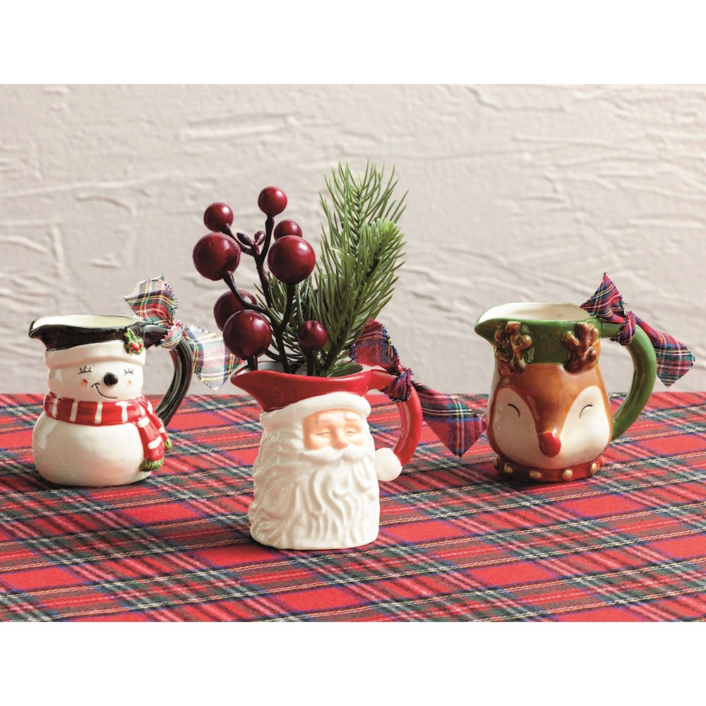 Three ceramic Christmas mini pitchers featuring a snowman, Santa, and reindeer on a plaid tablecloth.