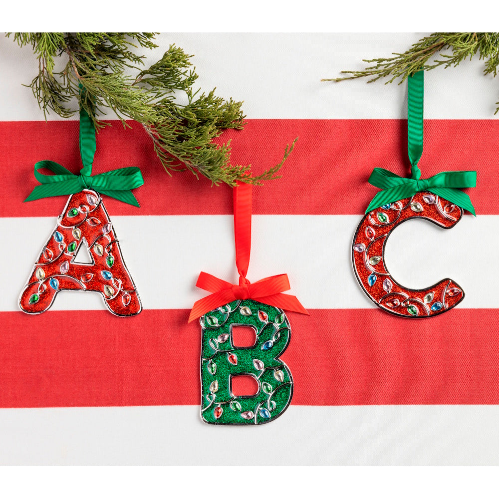 Sparkly Christmas alphabet ornaments A, B, and C hanging against a red and white striped background with pine needles.