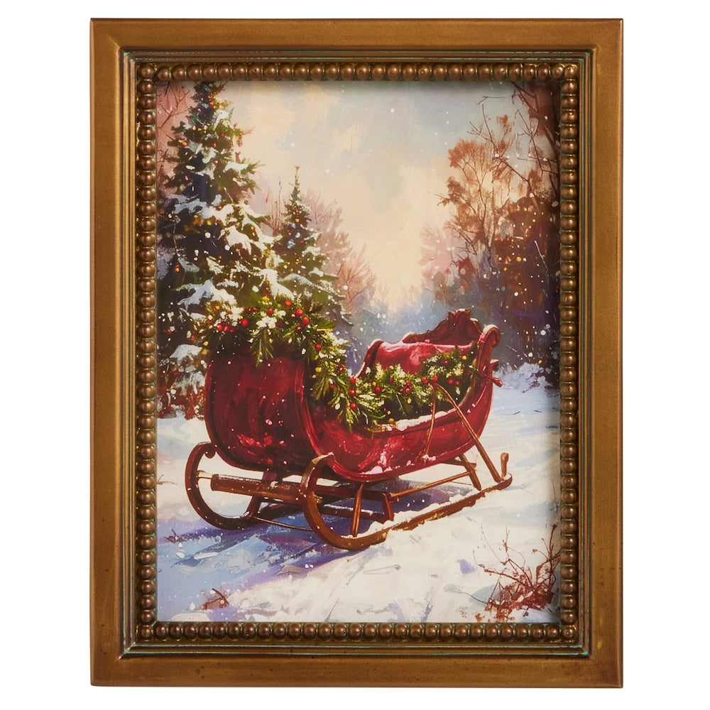 Framed Christmas wall art showing a red sleigh filled with garland parked in a snowy forest.