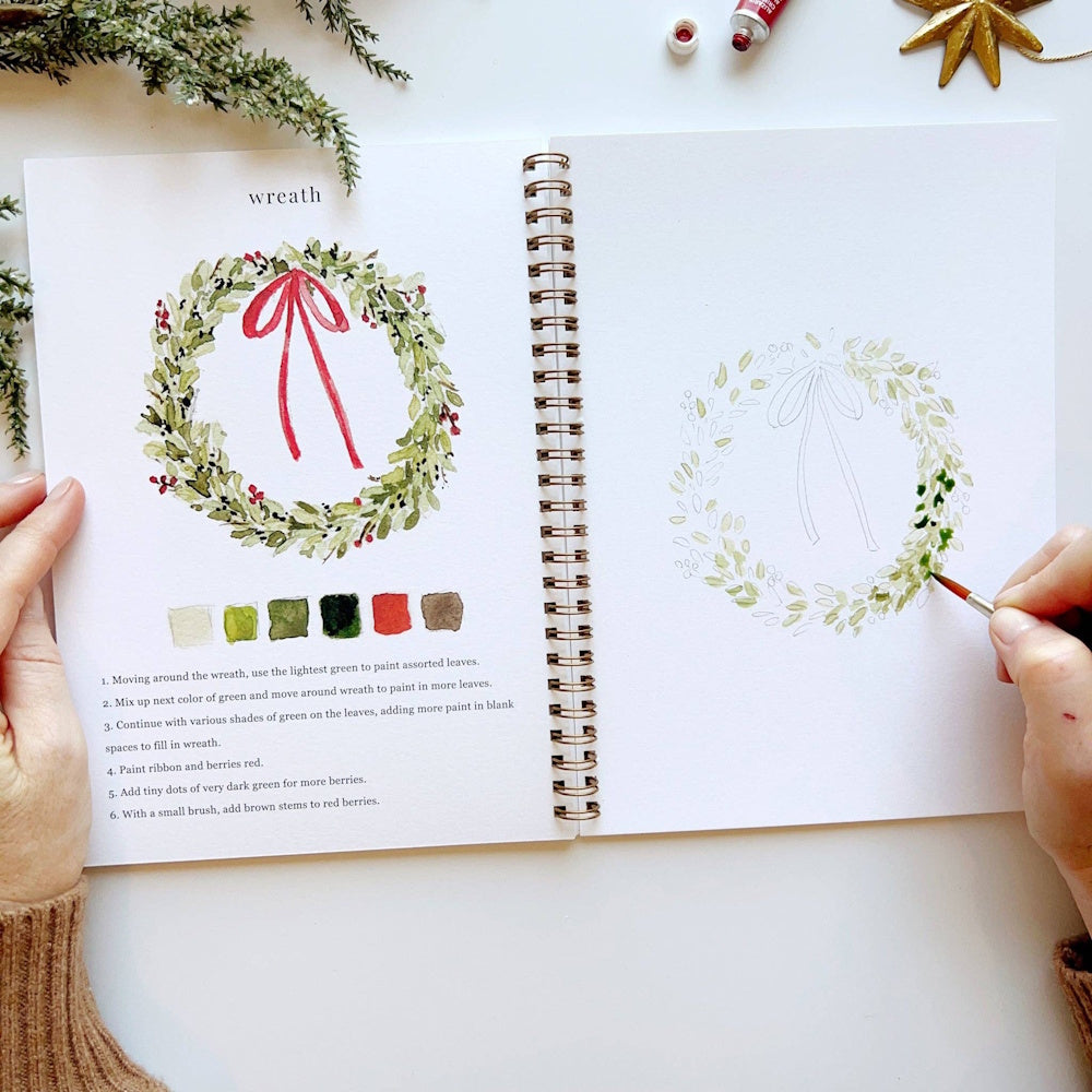 Top-down view of hands holding a Christmas watercolor workbook while painting green wreath leaves.