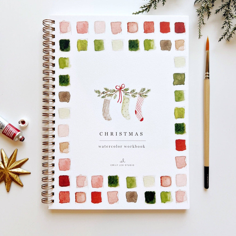 Christmas watercolor workbook with paint swatches, a paintbrush, and watercolor tube beside it.