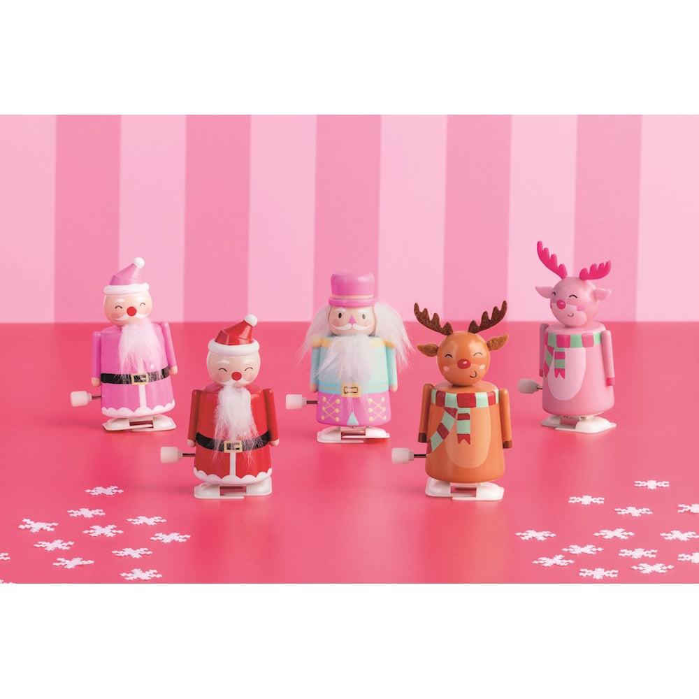 Five Christmas wind-up toys, including Santas, a nutcracker, and reindeer, against a pink backdrop.