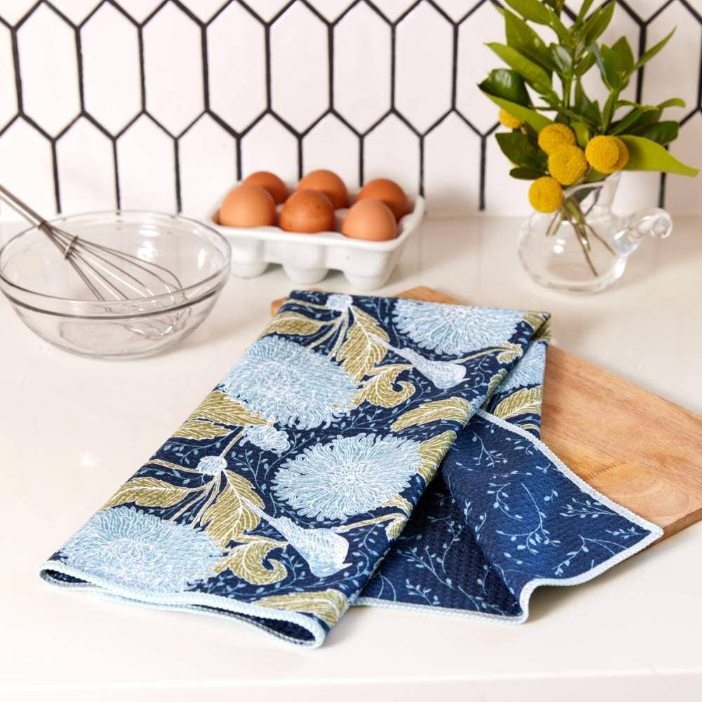 Blue and green floral tea towel draped on a white counter with eggs, whisk, and yellow flowers in a kitchen setting.