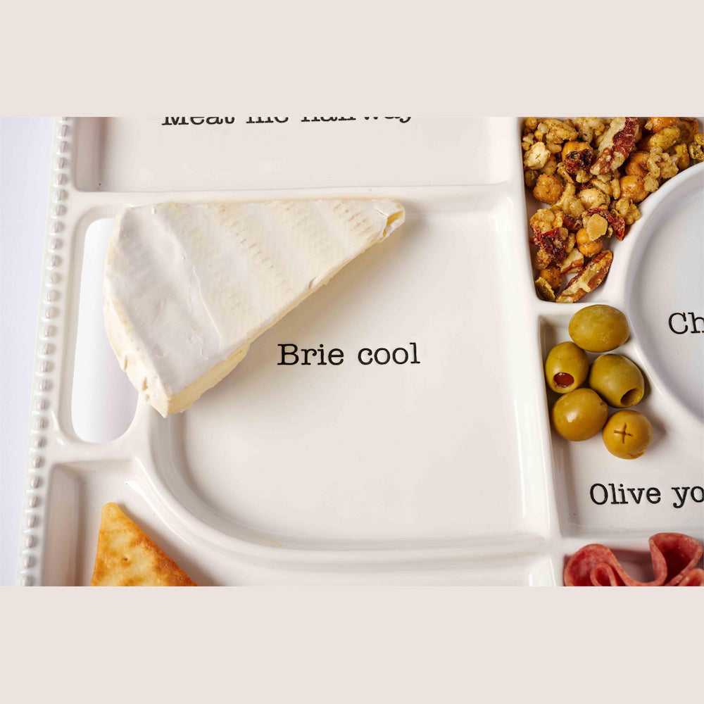 White ceramic divided charcuterie tray featuring brie cheese, olives, and snacks with playful pun labels.