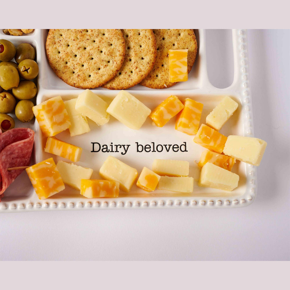 White ceramic charcuterie tray with "Dairy beloved" text, featuring cubed cheddar cheese, crackers, green olives, and salami.