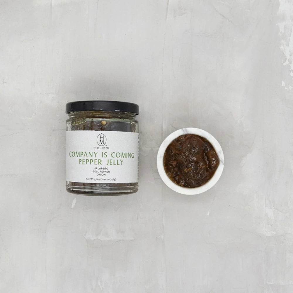 A 9-ounce jar of Hazel Mazel "Company Is Coming" Pepper Jelly next to a small white bowl filled with the dark jelly.