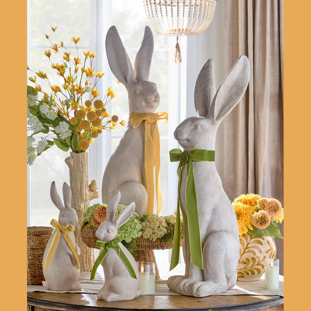 Four stone bunny statues with colorful ribbon bows sit on a table amidst vibrant floral arrangements. The room feels lively and festive.