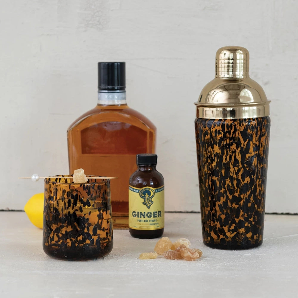 Amber tortoise shell cocktail glass and shaker set with ginger syrup and bourbon bottle on a white background.