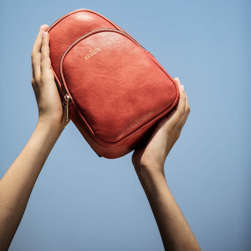 Two hands holding up a terracotta-colored Kedzie backpack against a clear blue sky.