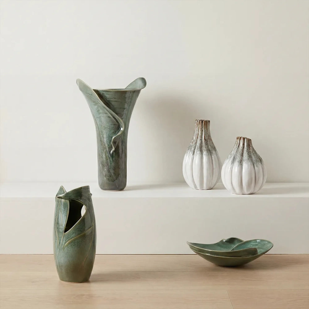 Five ceramic vessels in shades of green and white sit on a minimal, multi-tiered white shelving unit.