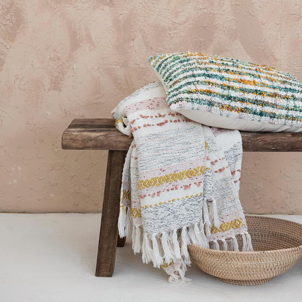 Boho textured throw blanket and striped lumbar pillow on a rustic wooden bench with a woven basket.