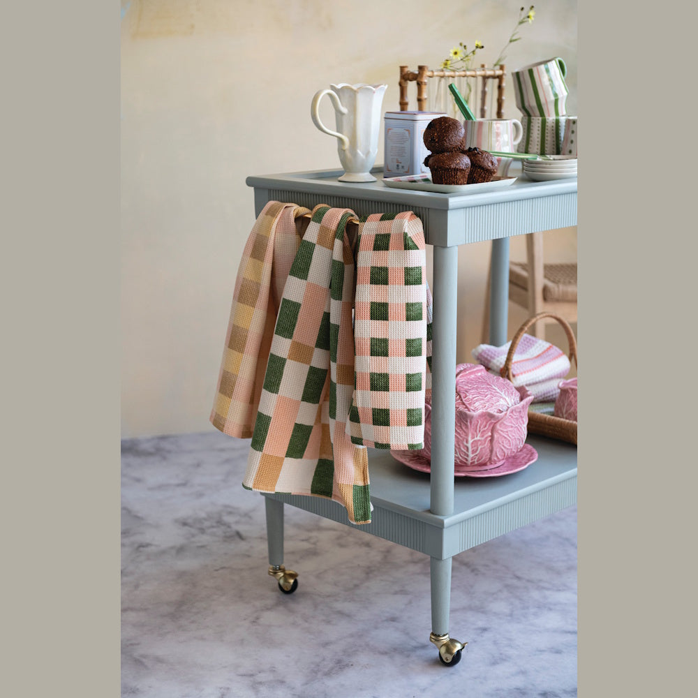 Plaid waffle weave tea towels in green and pink hanging on a light blue kitchen cart with muffins and tea accessories.