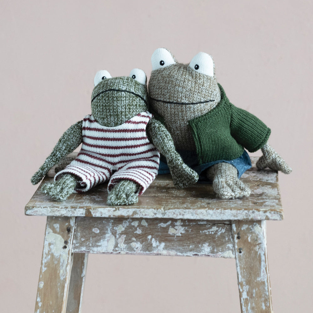 Two knit plush frogs wearing outfits sit side-by-side on a weathered wooden stool against a pink background.