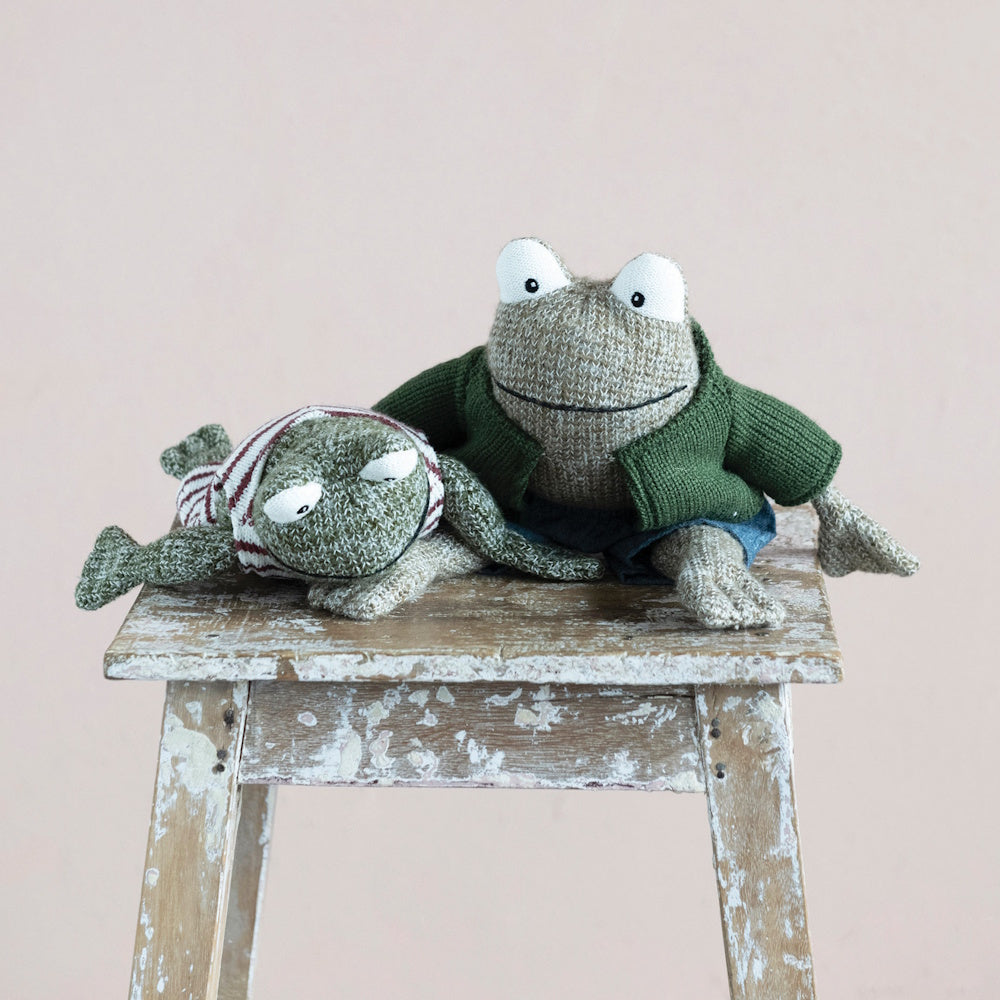 Two knitted toy frogs wearing outfits sit on a rustic wooden stool against a plain light background.