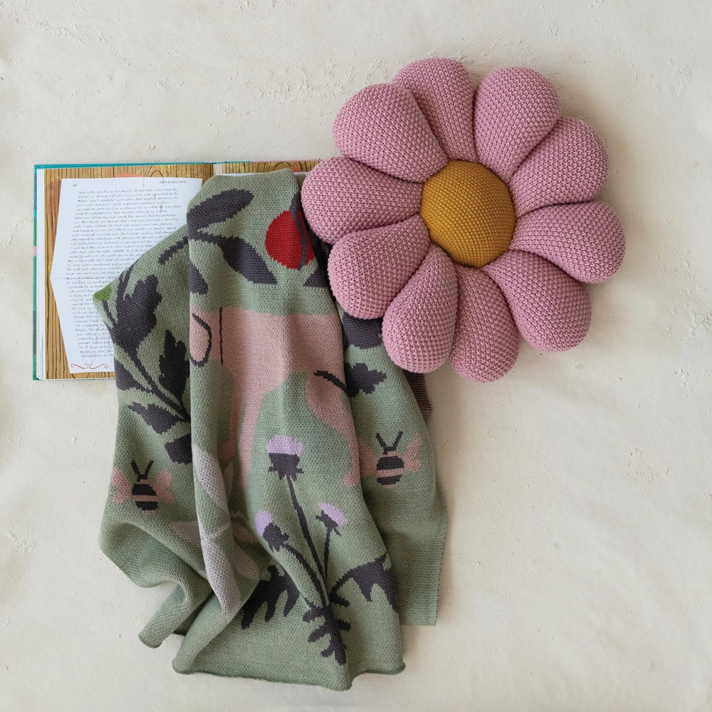 Green botanical knit baby blanket with bee and deer motifs next to a pink flower-shaped pillow and an open book.