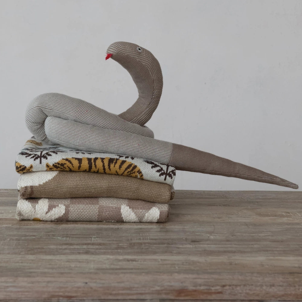 A knit snake plush coiled on a stack of patterned baby blankets, including tiger and daisy designs, on a wooden surface.