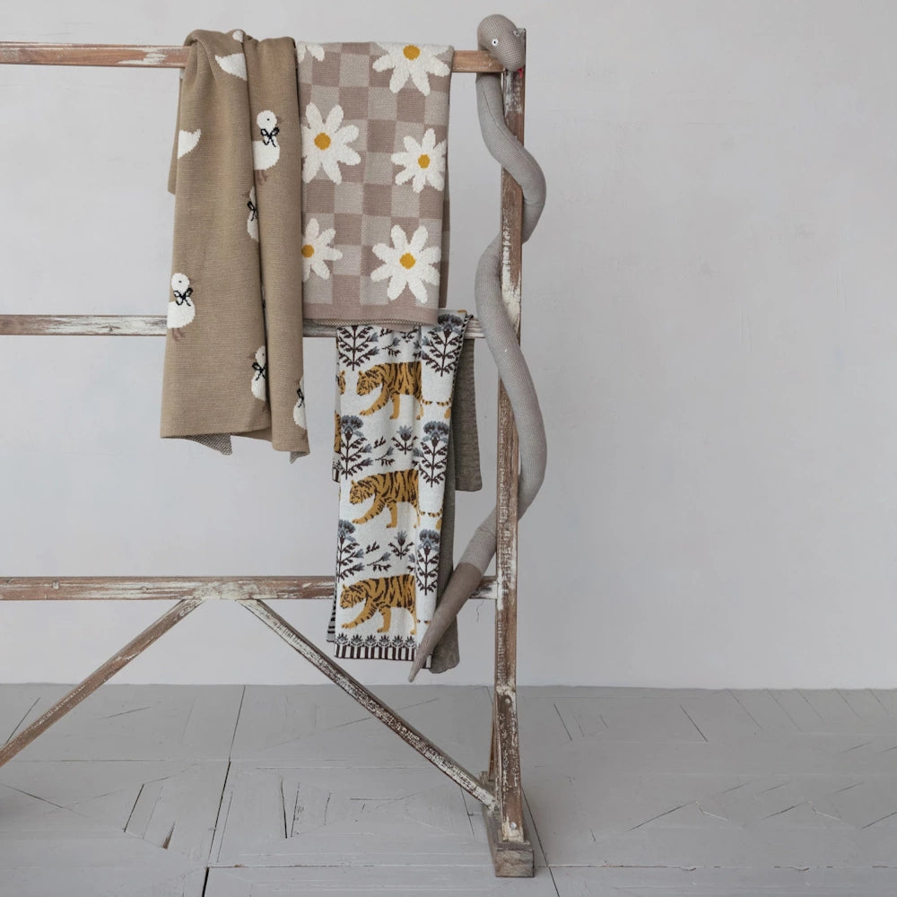 Knit baby blankets with daisy, tiger, and duck patterns hanging on a wooden ladder next to a plush snake toy.