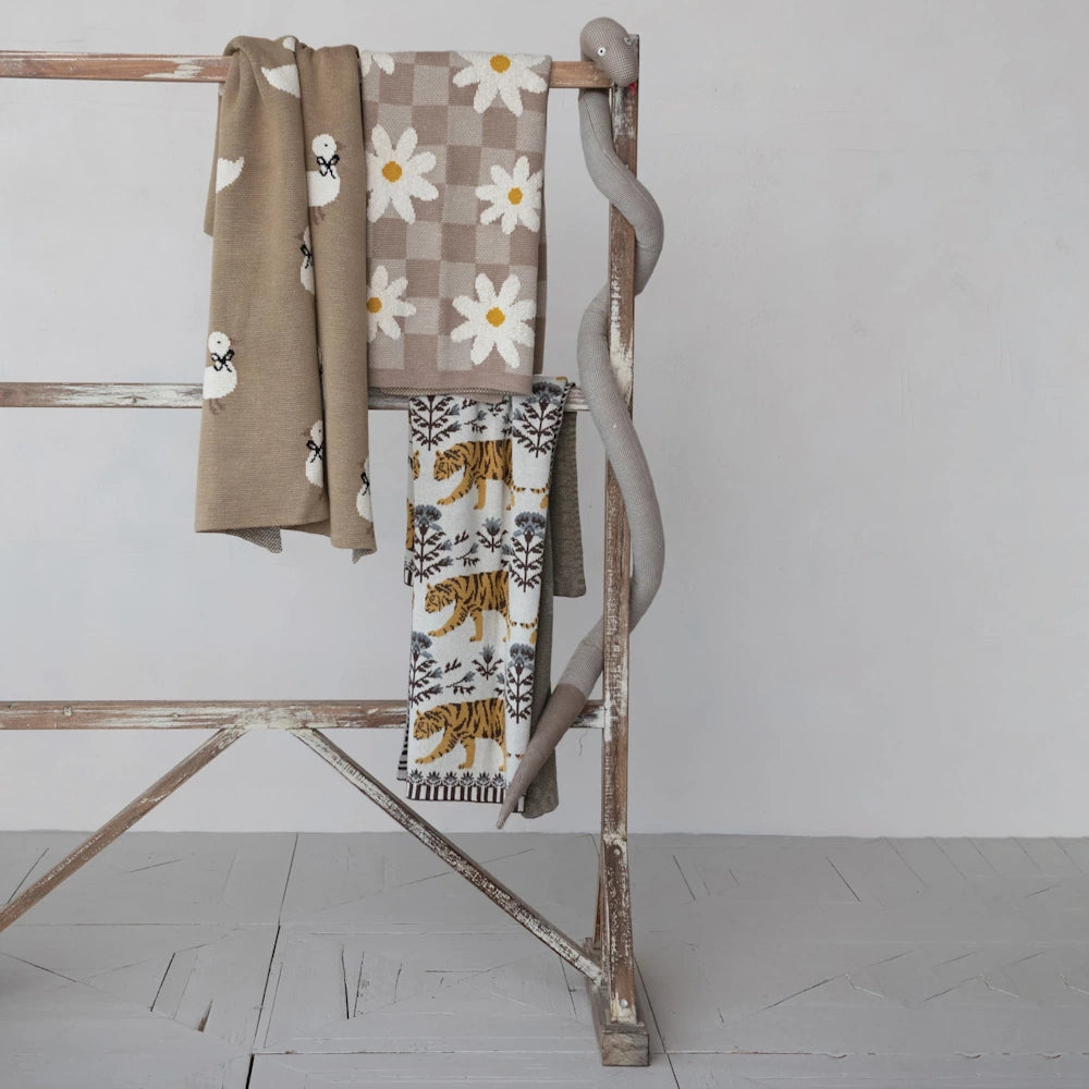 Knitted baby blankets with duckling, daisy, and tiger patterns hanging on a wooden ladder with a plush snake toy.