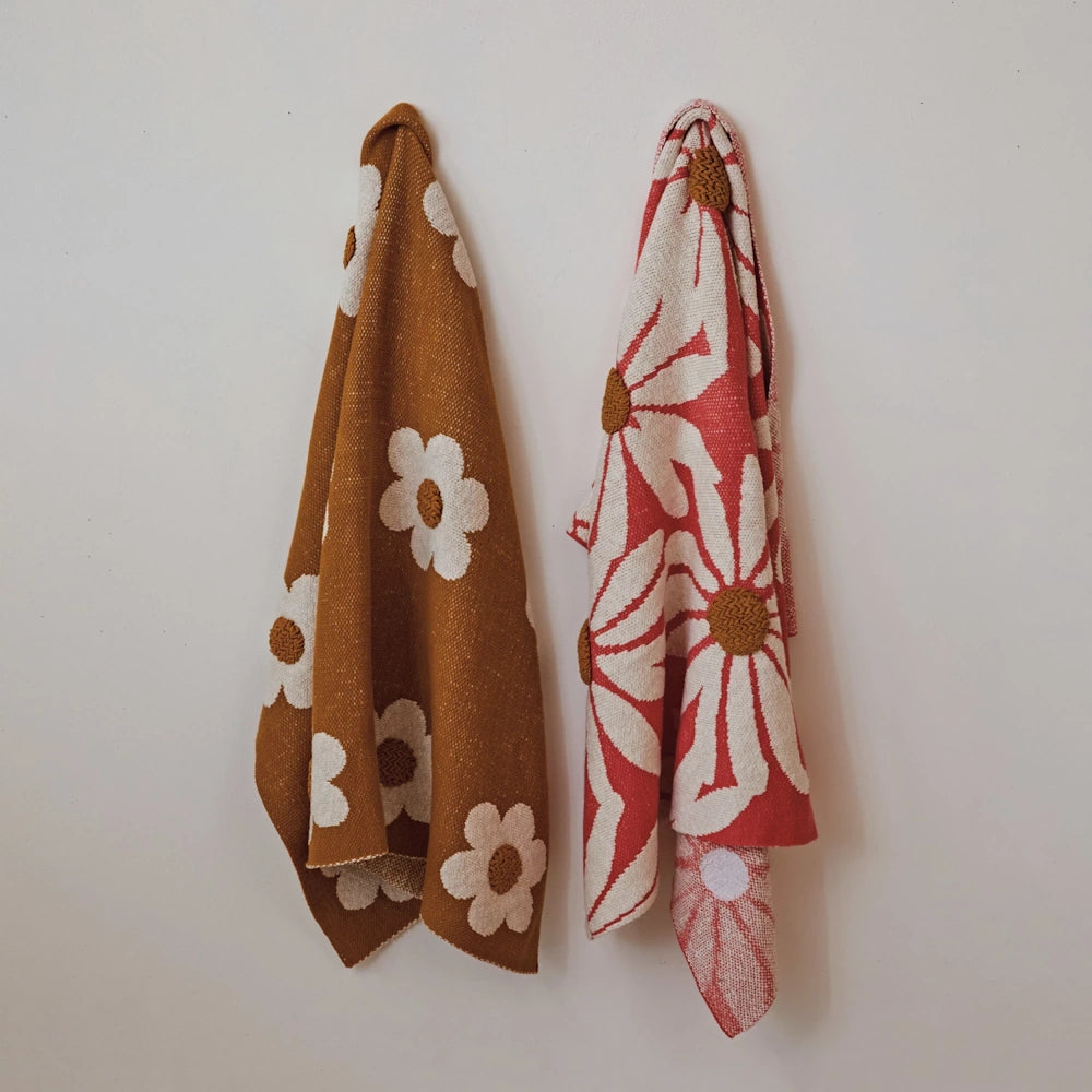 Two retro floral patterned blankets, one mustard yellow and one coral pink, hanging against a white wall.