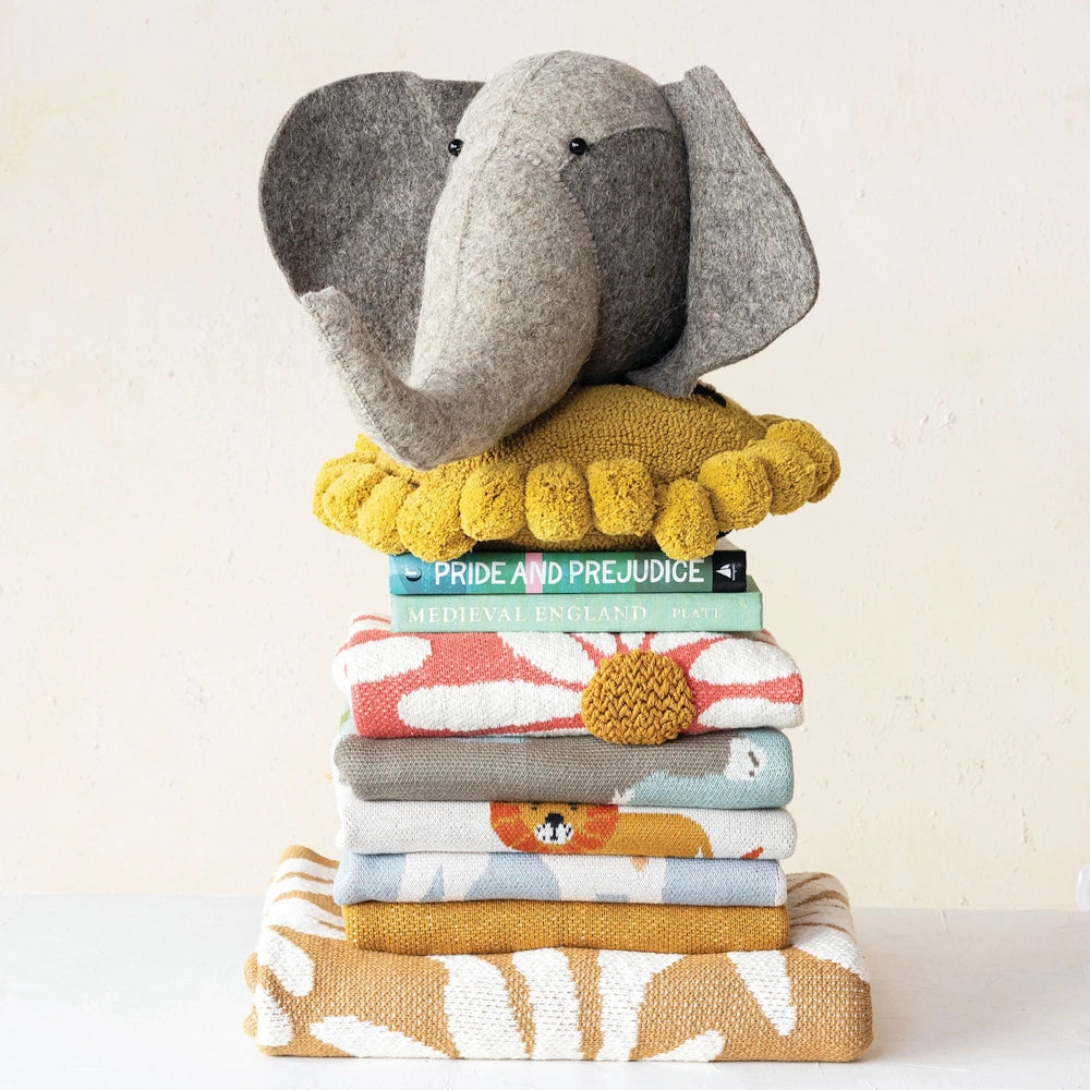 Felt elephant head wall decor resting on a yellow pom-pom pillow atop a stack of colorful patterned blankets and books.