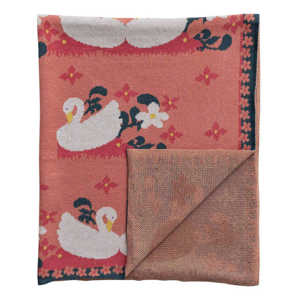 Folded peach cotton knit baby blanket featuring white swans, flowers, and a decorative floral border. Ideal nursery bedding.