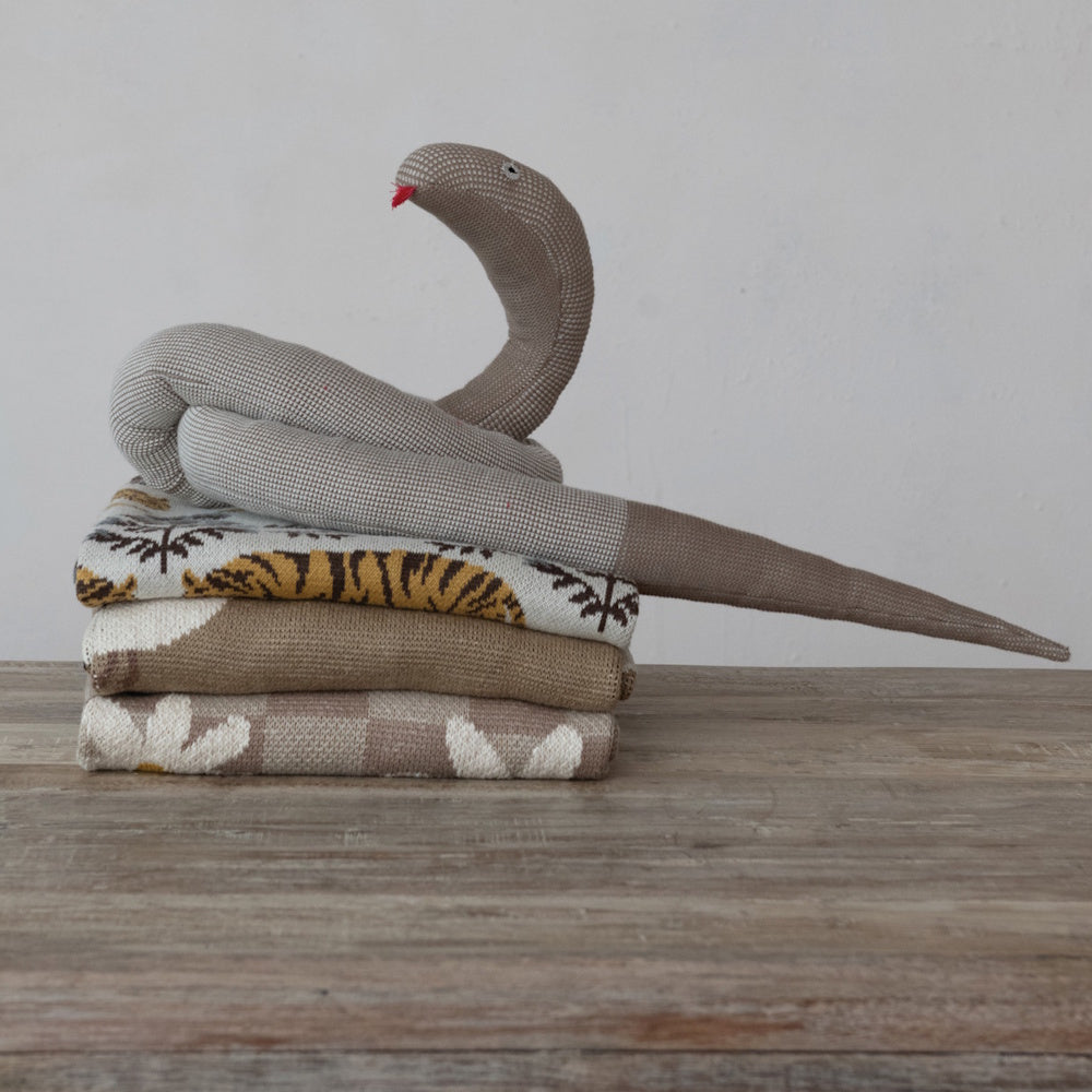 Knitted snake plush toy coiled on a stack of tiger and floral pattern throw blankets on a rustic wooden table.