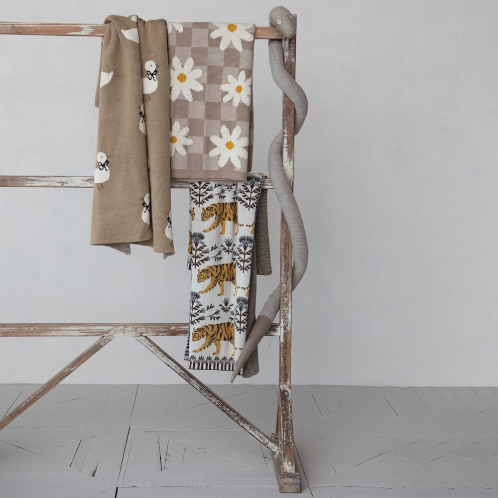 Knitted nursery blankets with daisy, tiger, and swan patterns hanging on a wooden ladder with a plush snake toy.
