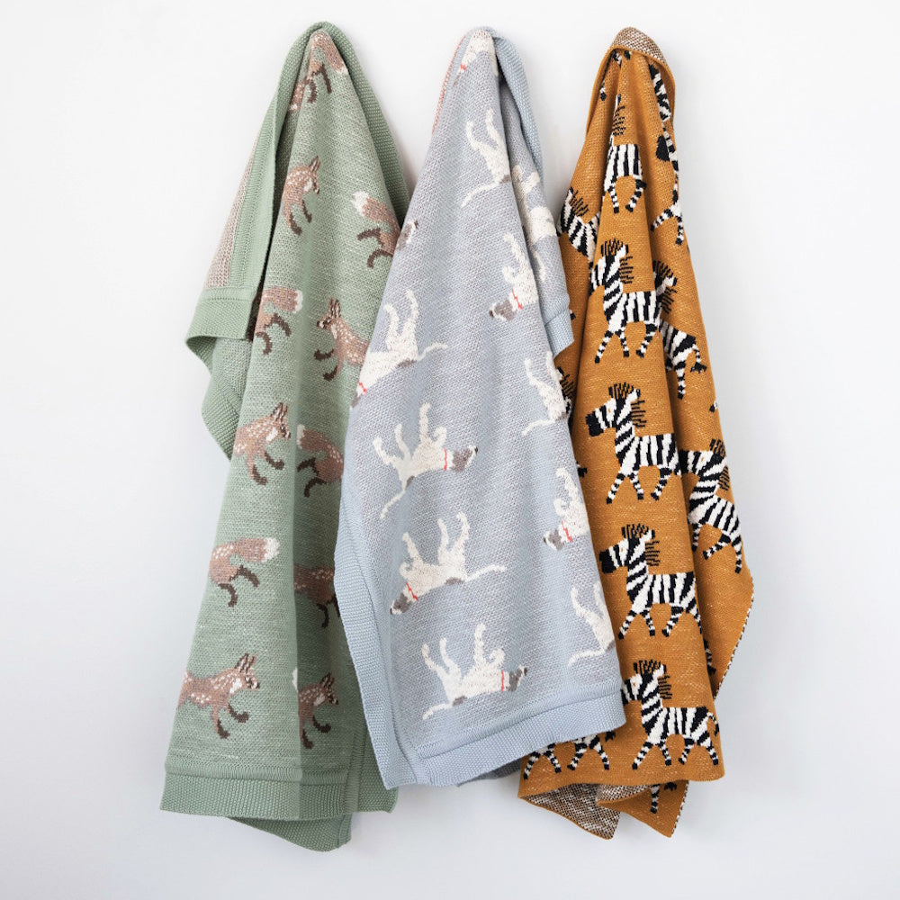 Three cotton-knit baby blankets hanging on a wall, featuring green fox, grey dog, and yellow zebra patterns.