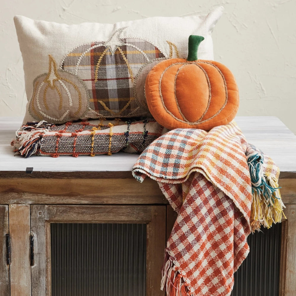 Beige lumbar pillow with appliquéd pumpkins, an orange plush pumpkin, and plaid throws on a cabinet.