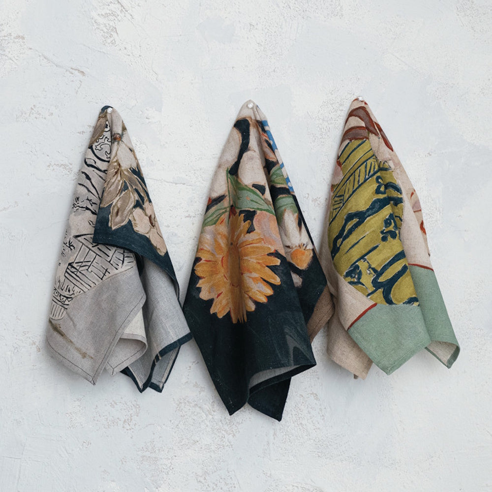 Three patterned fabric tea towels with floral and vase designs hang pinned to a textured white wall.