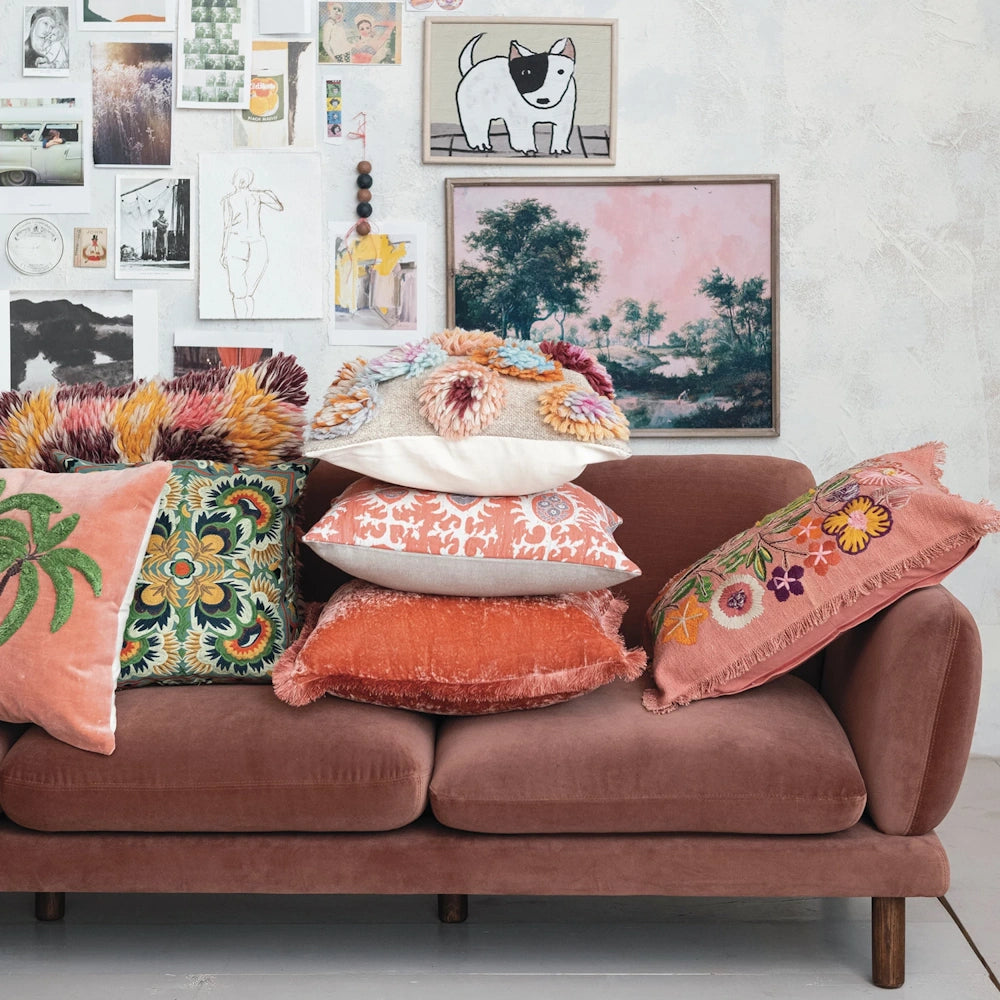 Colorful textured throw pillows stacked on a mauve velvet sofa against a gallery wall with art and photos.