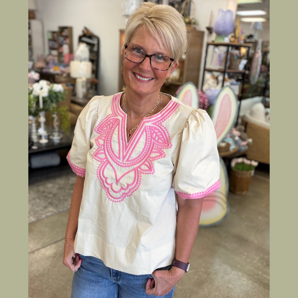 A smiling woman with blonde hair, glasses, and a cream-colored top with pink embroidery stands in a store.