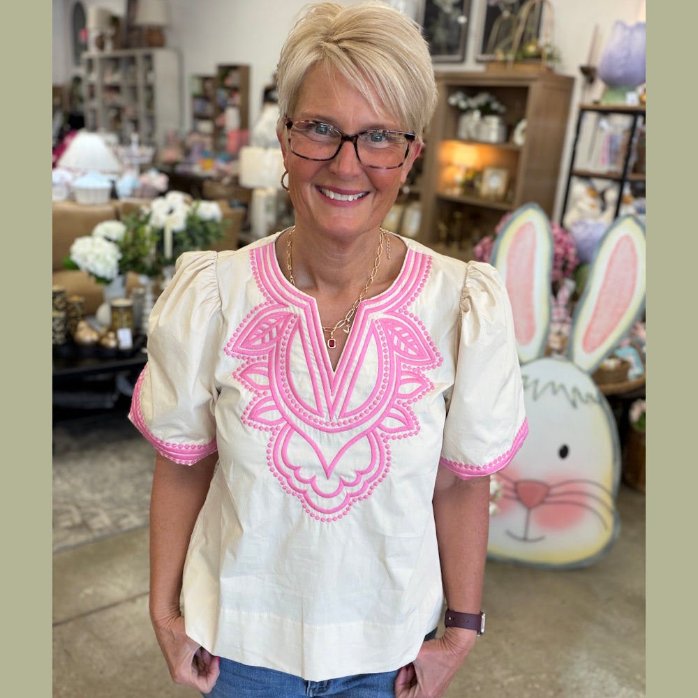 Smiling woman wearing a cream cotton poplin top with bright pink embroidered details around the neckline.