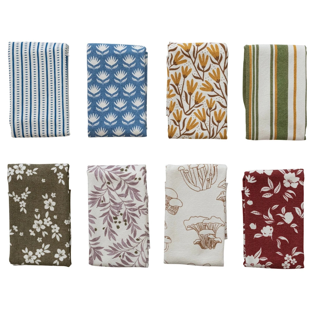 Assorted folded cotton terry cloth tea towels with botanical, striped, mushroom, and floral prints in earth tones.