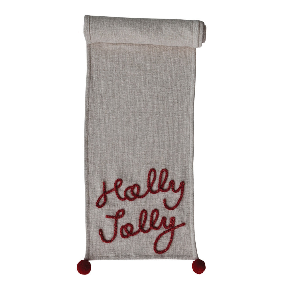 Cream woven table runner embroidered with red "Holly Jolly" script and accent red pom-poms.