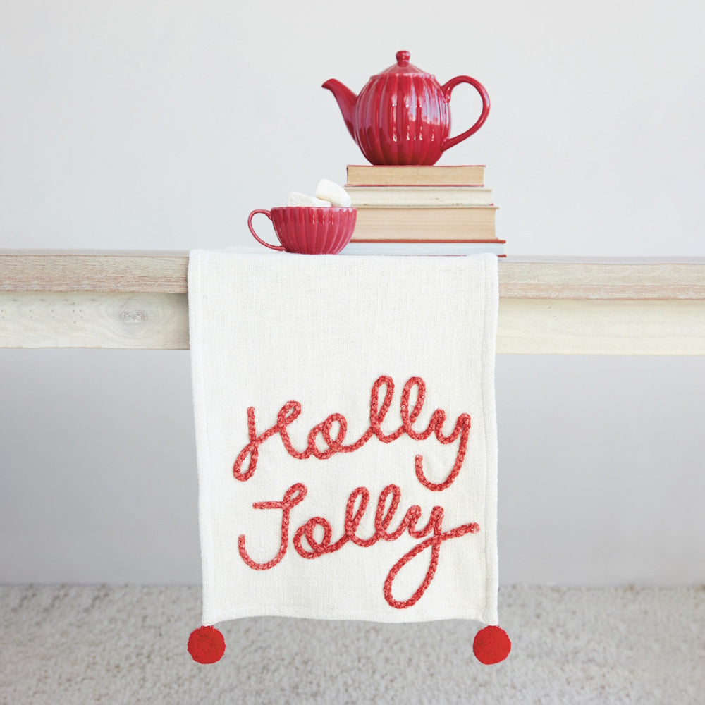 Cream table runner with red "Holly Jolly" embroidery, red pom-poms, and a red tea set.