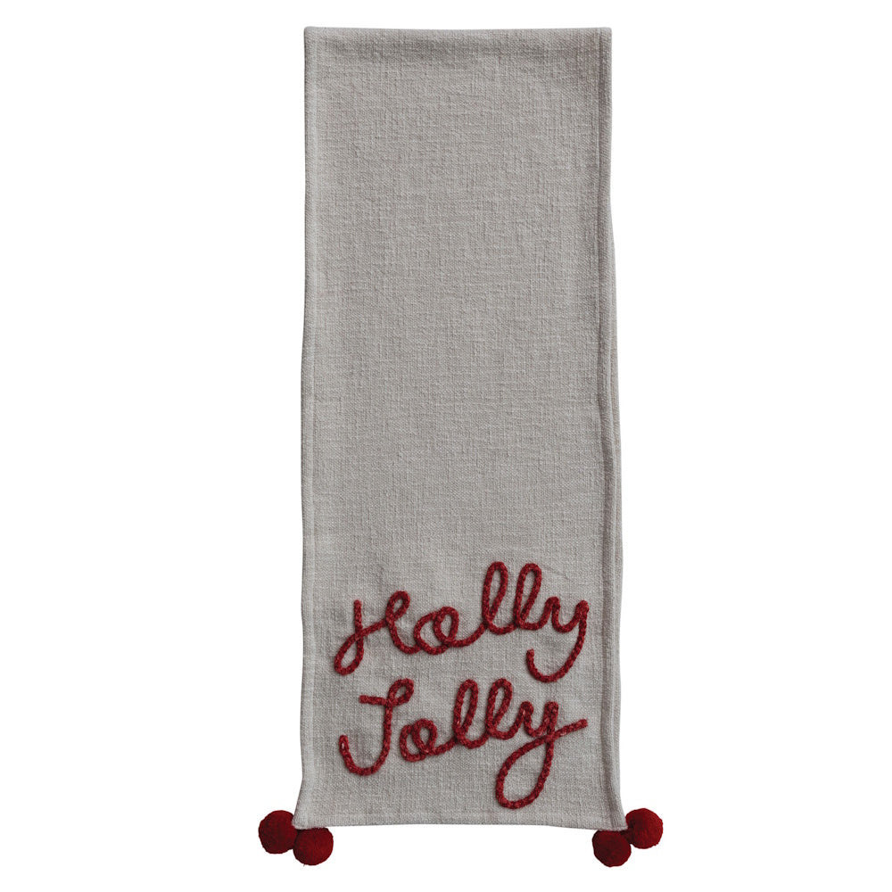 Grey table runner with red embroidered "Holly Jolly" text and corner pom-pom accents.