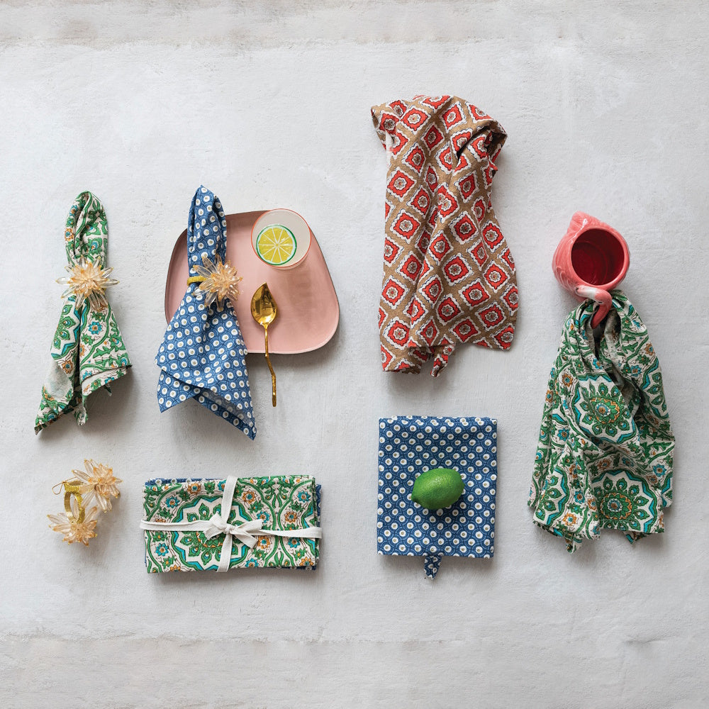 Patterned cotton slub tea towels in green, blue, and red block prints arranged with a pink plate and gold spoon.