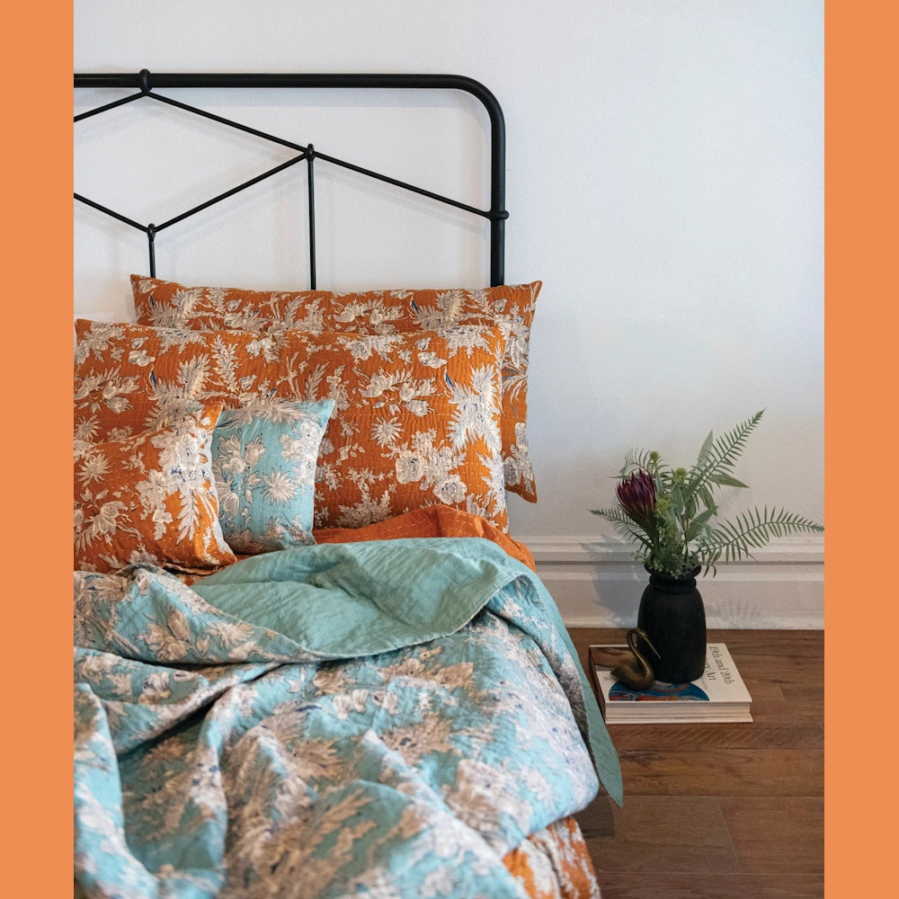 A bed with a black frame features patterned orange and teal bedding. A floral arrangement sits on stacked books.