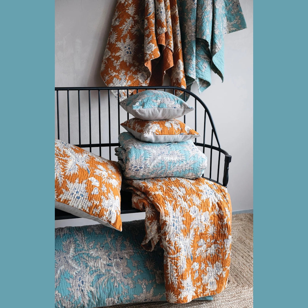 Floral printed cotton voile bed cover and queen shams in orange and teal, stacked on a black metal bench.