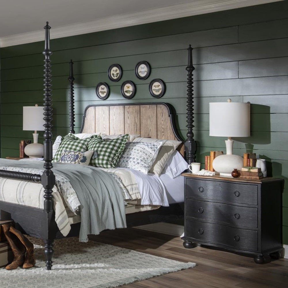 Bedroom with black three drawer accent chest used as a nightstand beside a black poster bed and lamp.