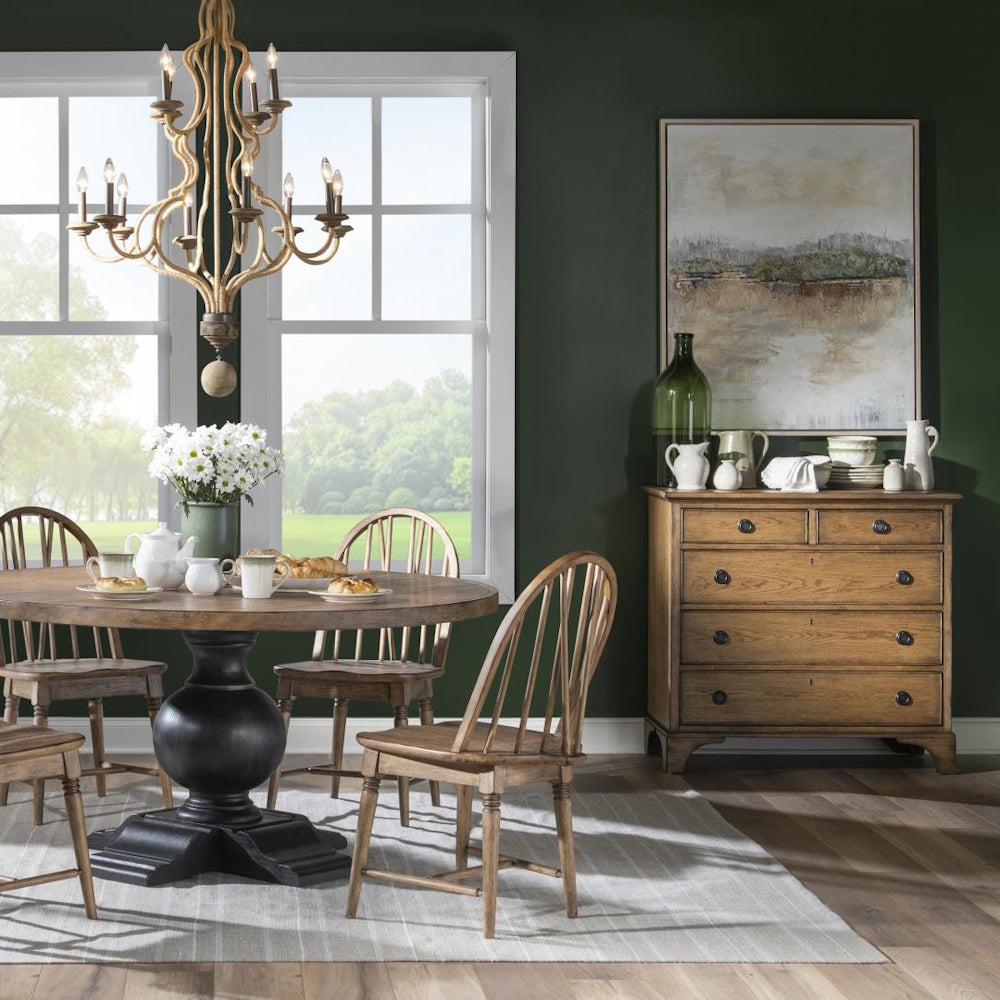 Dining room with rustic wood accent chest, round table, spindle chairs and dark green walls.