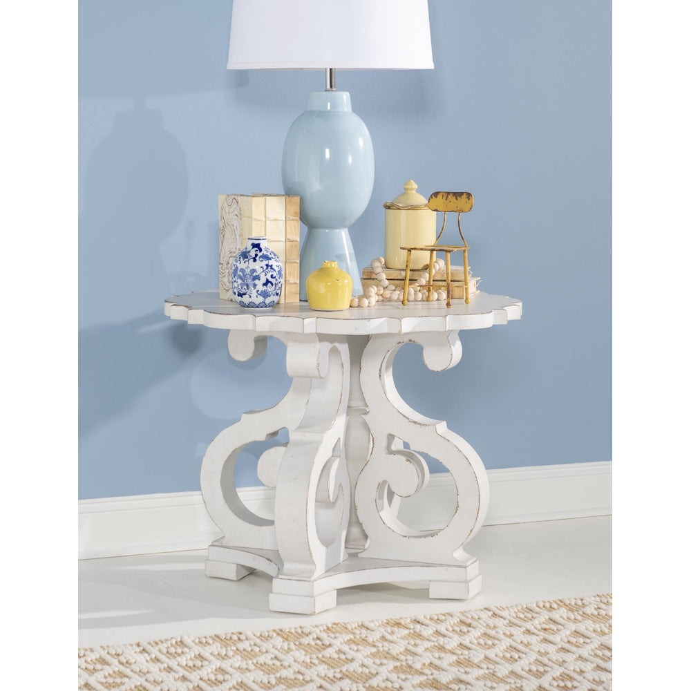 White distressed end table with ornate scrollwork base, styled with lamp, books, and decorative accents.