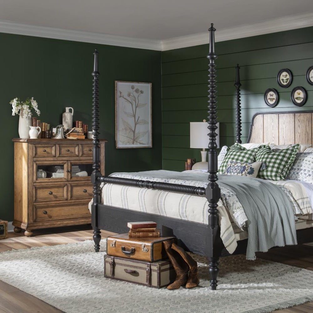 Rustic bedroom with black poster bed, wooden chest, green paneled walls, and vintage-style decor.