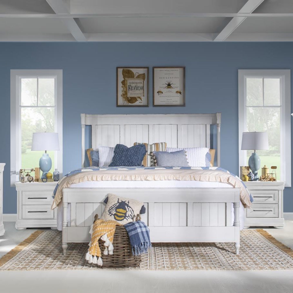 White paneled queen bed styled with blue and yellow bedding, flanked by matching nightstands and lamps.