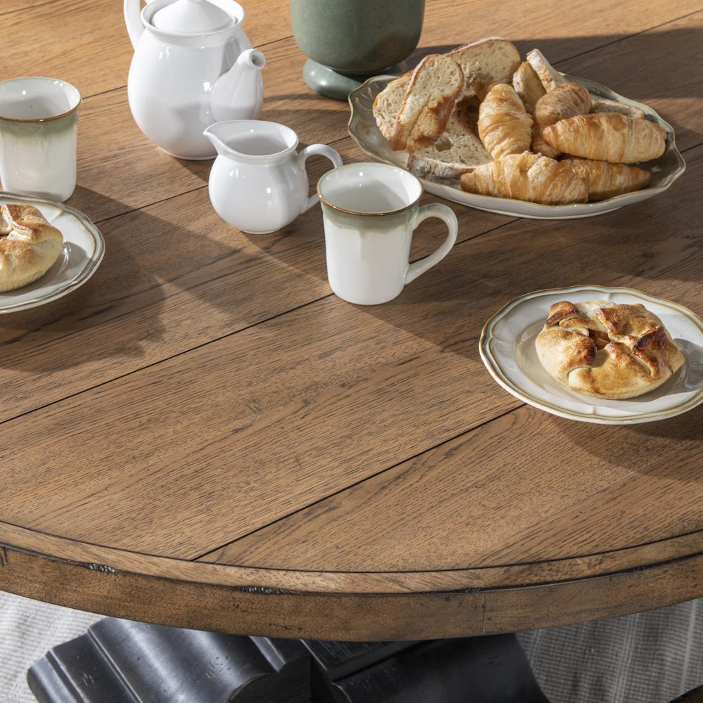 Close-up of a round wooden dining table set with pastries, a teapot, and mugs.