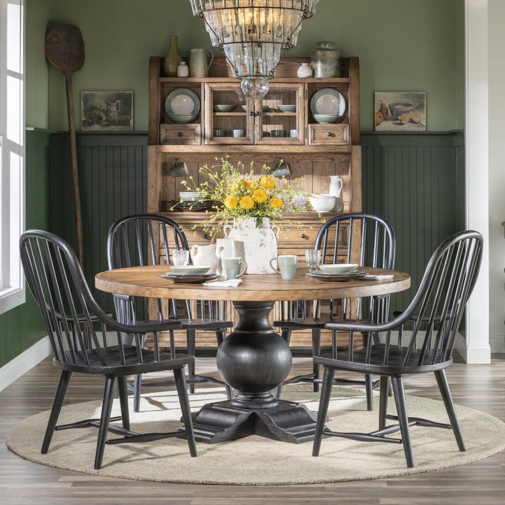 Rustic round wooden dining table with a black pedestal base and four black chairs in a green room.