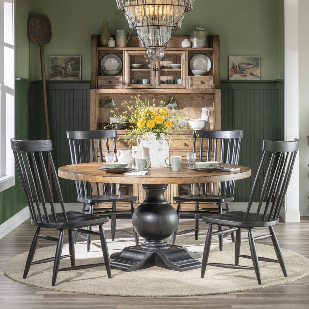 Round wooden dining table with a black pedestal base, surrounded by four black chairs in a green room.