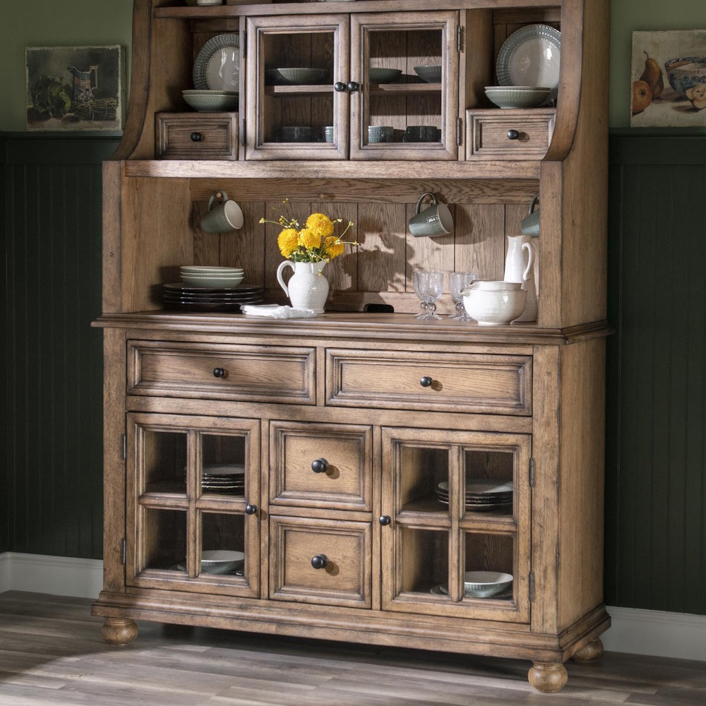 Rustic wooden hutch filled with dishes, mugs, and a white pitcher with yellow flowers against a dark green wall.